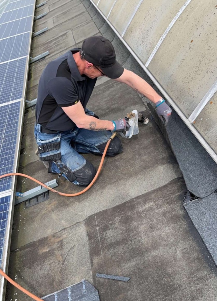 Ben van Eindhoven aan het werk op een plat dak naast zonnepanelen, met brander en dakbedekking